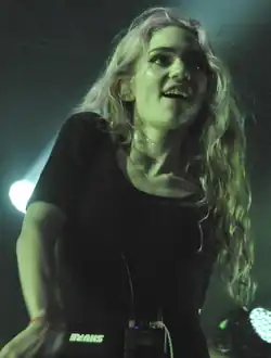 A woman with long, wavy, bleached hair wears a black shirt and looks excited, performing behind a microphone as pale green light illuminates her.