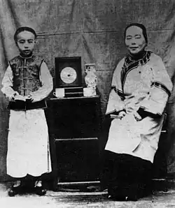 A black and white photo of a young boy and a partially seated older woman. Both are wearing traditional Chinese clothing and standing on either side of a cabinet holding pottery.