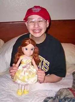 A young woman sitting upright in a bed, her legs under the blanket and the headboard and pillows behind her. She holds a large doll in front of her and is wearing a T-shirt, large glasses and a red baseball cap while smiling broadly