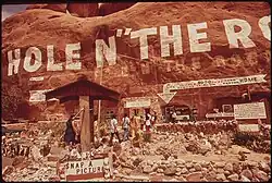 very large painted sign on the side of a sandstone cliff reading Hole N" The Rock with arrows pointing to the entrance. A handful of visitors are visible walking towards the door. Multiple signs are visible as well as a small rock garden.