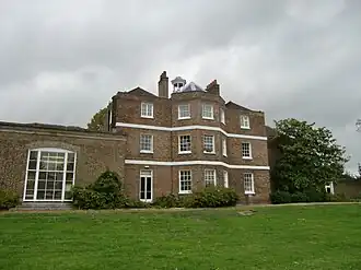 A three-storey brown brick building with a cupola, and a single storey extension on the left, the foreground is a green lawn