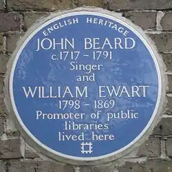 A Blue plaque on a brick wall with the words "John Beard C1717&nbsp;– 1791 Singer and William Ewart 1798&nbsp;– 1861 Promoter of Public Libraries
