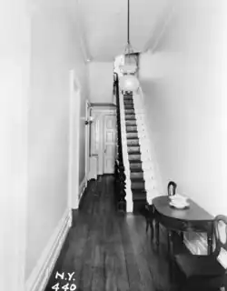 A black-and-white image of the house's first-floor hall. There is a narrow wooden staircase on the right, leading to the second floor. A chandelier hangs from the ceiling, and there is some wooden furniture at the bottom of the staircase.