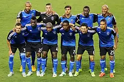 Eleven soccer players wearing blue jerseys and black shorts standing in two lines posing for a pre-match photo.