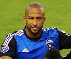 A man with a shaven head wearing a blue soccer jersey.