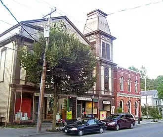 A large building with brown peaked roofs and stucco and half-timber face. There is a brick building next to it on a street corner.