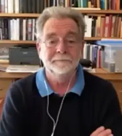 blurry image of Hugh White wearing a dark blue sweater over a light blue shirt, appearing to sit in front of a webcam, with white earphones, arms crossed, gaze directed slightly below camera