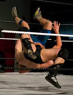 Behind ropes, only legs and a hand are visible of two grappling men.