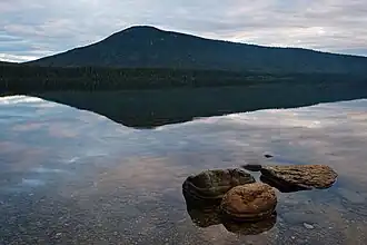 A calm lake reflects a partially clouded sky. A cluster of rocks protrudes from the water at the shore near the viewer, while across the lake stands a wide, tree-covered mountain with around peak.