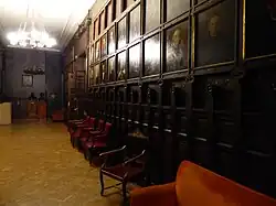 Photograph of a room. The wall is covered in portraits of the same size and dark wood. There are old-style chairs.