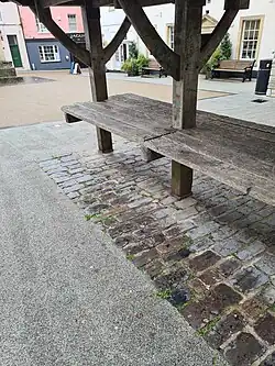 Colour photograph of the restored Shambles in the Market Place at Shepton Mallet. The bench and curved roof trusses are made of oak, and the floor is made of local stone and is brown in colour. The public library can be seen in the background.