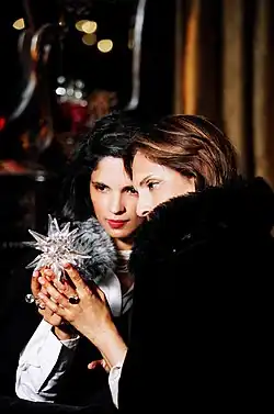 Two women from the left profile. Their heads touch each other's. The woman on the right is wearing a dark cloth, which covers her mouth area. She has brownish hair and with her left hand, holds a crystal star. The woman on the left is wearing a white shirt with a black cape around her and is also holding the star, with her right hand. Both women are looking at the star.