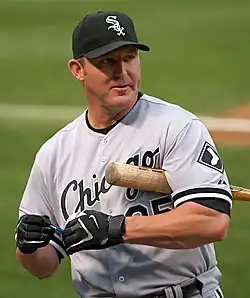 A man in a grey baseball uniform with "Chicago" on his chest in black with a black cap, black batting gloves, and a baseball bat under his arm.