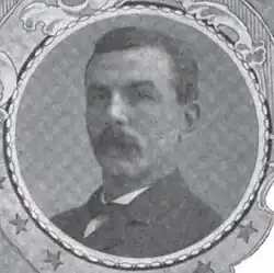 Portrait of a man with a drooping mustache and a dark suit coat, in a circular frame.