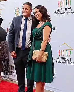 John Steenhuisen is on the left, wearing a black suit jacket and pants, with a white shirt and blue tie. Beside, on the right is Terry Steenhuisen, who is wearing a green sleeveless dress. They are standing on the red carpet posing. Behind them is a backdrop of text that says 'Opening of Parliament Address' with a symbol representing the front of the parliament building. The logo for the South African Parliament can be seen just behind John.