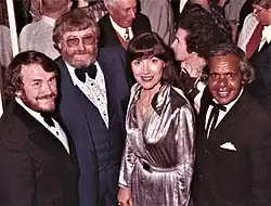 an indoors colour photograph with three white people and one Aboriginal man in formal attire in the foreground with a large gathering in the background