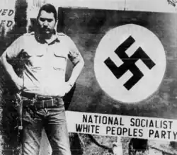 Black and white photo of Tommasi wearing a swastika and standing in front of a sign with a large swastika, which reads National Socialist White People's Party