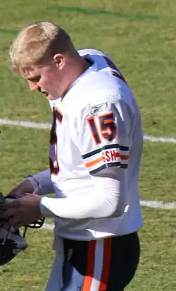 A photo of a helmetless football player in a white #15 jersey holding his helmet