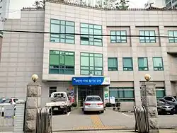 A three storey tall grey building with a blue sign above the door, which has Korean text written on it