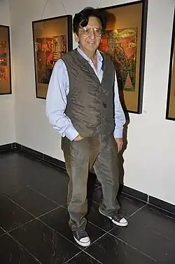 Kailash Surendranath wearing a navy blue suit and white shirt, standing against a marble-textured background, smiling confidently.
