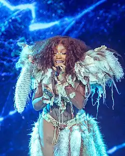 SZA kneeling onstage in front of a stage screen showing a blue linear patterns