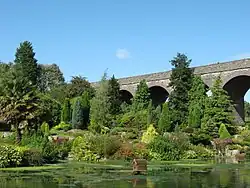 View of a garden containing a number of evergreen trees and a palm tree, with a lake in the foreground and the arches of a disused railway viaduct in the background.