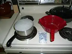 Kraft Macaroni & Cheese preparation—pot of boiling water with pasta inside, package of cheese powder (labeled "Kraft cheese sauce mix"), and colander to separate the noodles from the cooking water