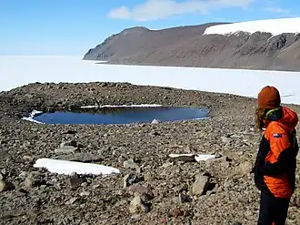 A rocky landscape with a small lake in the centre. An ice covered harbour is visible in the background with hills in the distance