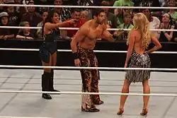 A brunette Caucasian woman and dark-haired Caucasian man in a wrestling ring, pointing and yelling at a blonde Caucasian woman with her back to the camera.