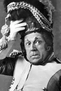 head and shoulders shot of middle-aged white man in 1840s French military uniform, with one hand steadying his unwieldy headgear and making a comically anxious face