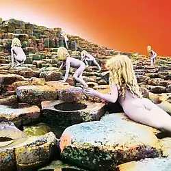 Six nude children with long blonde hair scramble up a stairstep series of basalt rocks ascending away from the viewer, with an orange-white sky above