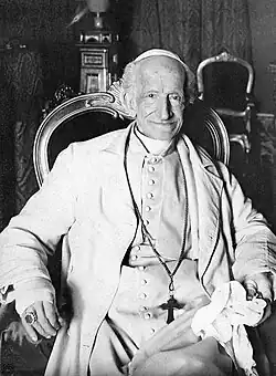 An elderly man wearing a white suit, a long cross-necklace, a ring, a white zucchetto, and a large ring sitting in an ornate chair and holding a cloth