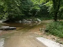 A color photograph depicting an improved ford across a small stream with a gravel road and lush foliage.