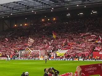 A stand which is full of people standing to support their team. There are a number of flags, scarfs, and banners in the crowd.