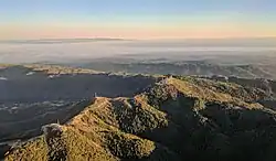 Loma Prieta (the peak just right of center) and other nearby peaks are decorated with television broadcast towers and other communication towers, serving the Santa Clara Valley. Fog-shrouded Monterey Bay and the Monterey peninsula are visible in the background in this late-afternoon approach to San José International Airport. On the left-near side you can see Crystal Peak and Mt. Chual.