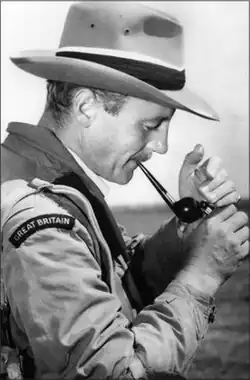 Swansea, a man in early middle age with neat, cropped hair, in his shooting attire: he wears a wide-brimmed hat on his head and a "Great Britain" flash on his shoulder, and is lighting a pipe to smoke.