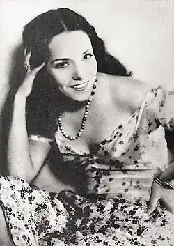 Black and white portrait photograph of Lupe Vélez from 1930, showing her leaning forward with her hand in her hair, wearing a patterned dress with exposed shoulders