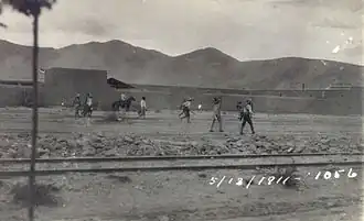 A group of men wearing sombreros walking along a dirt road between a railroad track and a line of buildings. Dated 5/13/1911.