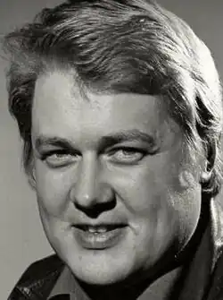 Publicity shot of half-smiling, clean-shaven man, with strong features and short, swept-back hair.