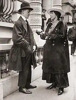 Mary Newcomb is standing on sidewalk selling tickets to a man with a hand in his pocket getting money out