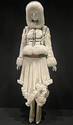 Ensemble in cream with brown accents. Top is in fur, with hood and long sleeves. Bottom is a skirt with asymmetrical hem lined with puffs.