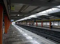 Picture of the Line 5 side platforms, where some people stands. The platforms are made of marble. In the far background, to the right, some cars are visible.