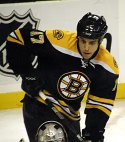 A frontal view of a hockey player in his early twenties, cut off at the thighs, during a hockey game. He is wearing a black and yellow jersey with a logo containing a capitalized "B". The number "17" is visible on his jersey's left sleeve. He is looking downwards with an intent expression on his face.