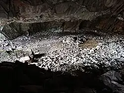 Photograph of the large "living room" from the small "bedroom" of "The Cave Hotel". The "living room" is on a lower level and is relatively flat, with a raised platform of rocks towards the rear. This was possibly to provide a drier area, as the floor is wet with dripping water towards the front of the cave. At the time of visiting (February 2023), there was a fire platform and crude log bench in the centre of the living area. There was a bit of a scramble up to the "bedroom" area, which has a separate entrance/exit.