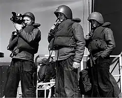 Sailors wearing Mark II talker helmets accompanying a photographer's mate capturing a bombardment by the USS Missouri (BB-63) in 1952