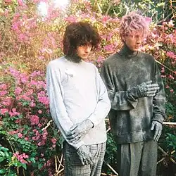 A brunette boy stands next to a man with pink hair in front of flowers. Both have cement on their hands and neck.