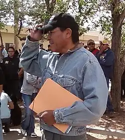 Mark Maryboy, a Navajo leader, wearing a cap and denim jacket, addresses a crowd during a public event while holding a folder.