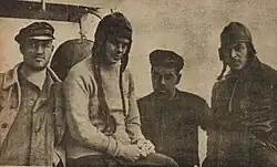 Photograph of a women wearing a hat with ear-flaps over long braids between one man on the left and two men on the right