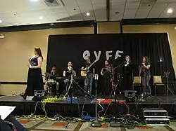 Photograph of several people on stage in front of a black curtain bearing the letters O V F F.