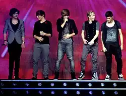 Five teenage boys standing next to each other on a stage and singing. From left to right: Harry Styles, Louis Tomlinson, Liam Payne, Niall Horan, and Zayn Malik.